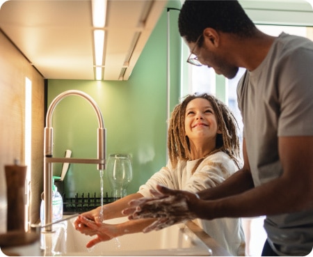 Homem em frente a uma pia de cozinha mostrando a uma criança como lavar as mãos corretamente como parte de uma rotina saudável de higiene pessoal.