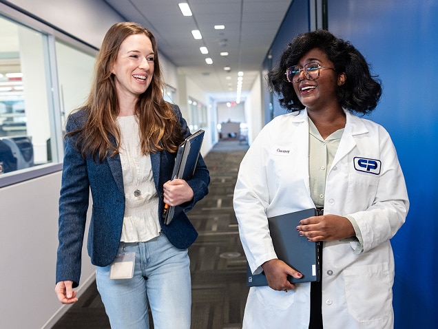 Deux collègues, dont une scientifique en blouse blanche, discutent en marchant dans le couloir moderne d'un bureau.