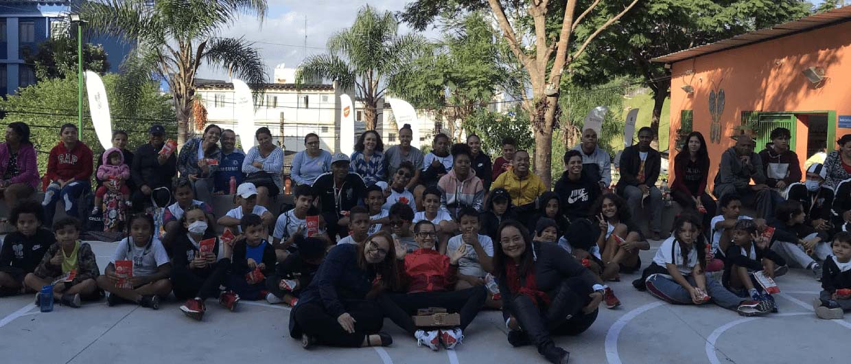 Grande grupo de crianças e adultos reunidos sentados em uma quadra externa durante um evento comunitário de dia ensolarado.