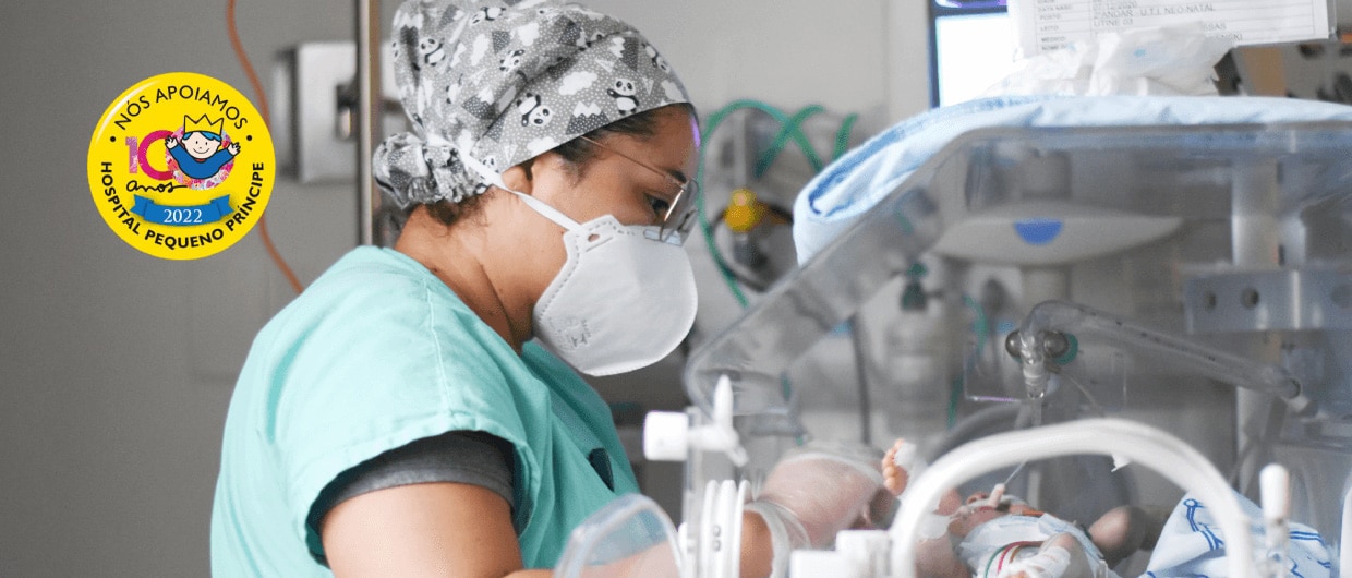 Médica de máscara e touca cirúrgica cuidando de bebê em incubadora neonatal no Hospital Pequeno Príncipe.