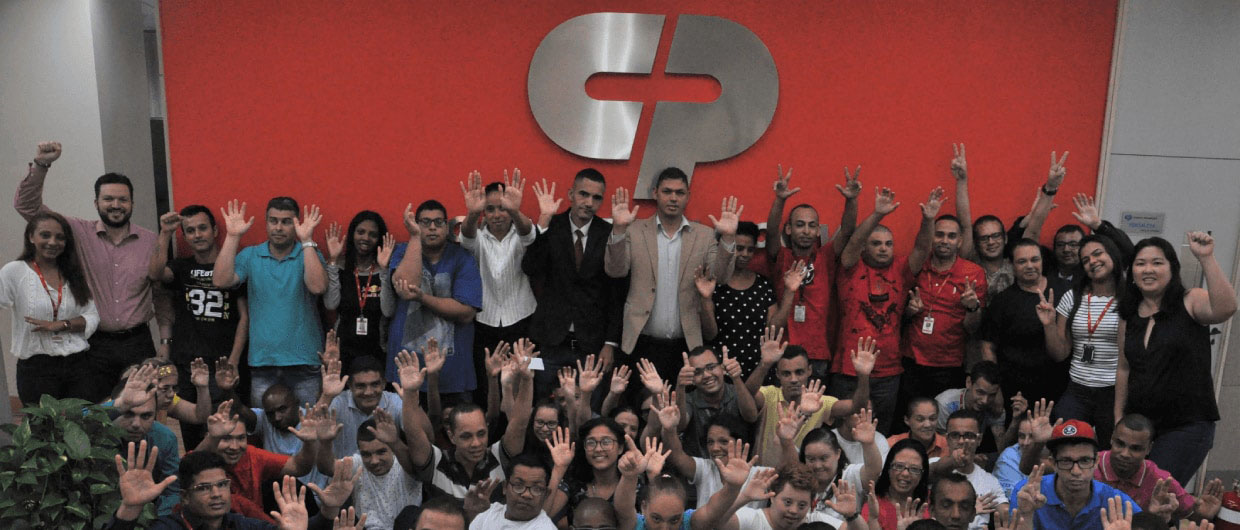 Grande grupo de colaboradores reunidos acenando em frente a uma parede vermelha com o logotipo metálico CP.
