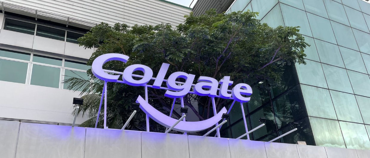Letreiro iluminado da marca Colgate com o ícone de sorriso instalado na fachada externa de um edifício corporativo moderno.