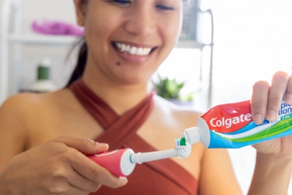 Mulher sorrindo enquanto coloca creme dental Colgate na escova de dentes em um banheiro.
